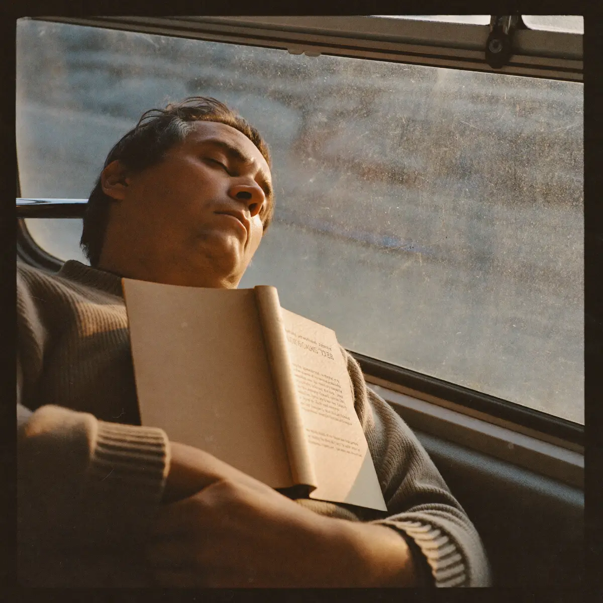 En man som sover mot en bussruta med en uppslagen bok på bröstet